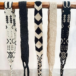 May include: Five woven fabric bands numbered one to five, hanging from a wooden rod. The bands feature various geometric patterns in black, white, and beige, each ending in braided cords with small tassels at the bottom of the textile strips.