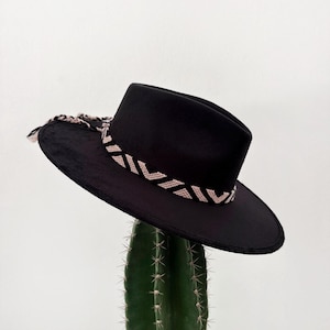 Puede incluir: Un sombrero negro de ala ancha con copa plana y ala rígida. El sombrero presenta una banda tejida decorativa con un patrón geométrico en beige claro y negro que envuelve la base de la copa, terminando en dos borlas trenzadas en el lateral.