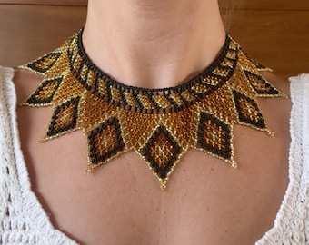 May include: A beaded collar necklace featuring a geometric pattern of black, gold, and bronze seed beads. The design consists of a dense, woven band at the neck with a series of pointed, diamond-shaped pendants extending downward in a sunburst style.