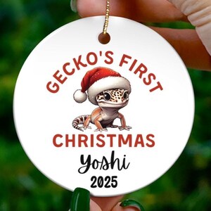Peut inclure: Une boule de Noël en céramique blanche avec une ficelle dorée, représentant un gecko portant un bonnet de Père Noël. Le texte indique "Gecko's First Christmas Yoshi 2025" en rouge. La boule est tenue devant un sapin de Noël flou.