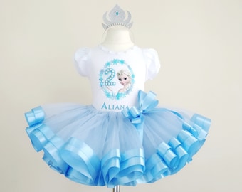 Peut inclure: Un tutu en tulle bleu clair avec un t-shirt blanc représentant un personnage de La Reine des Neiges et le chiffre 2. Le t-shirt porte également le nom "Aliana". Une couronne argentée avec des pierres bleues est posée sur le mannequin.