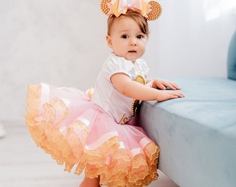 May include: A baby wearing a white top and a pink tutu with gold trim. The baby is also wearing a headband with gold ears and a gold bow. The baby is wearing pink shoes and is leaning against a blue surface.