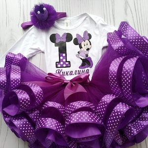 Può includere: Tutu di tulle viola con nastro a pois bianchi, un body bianco con un numero uno viola e una grafica di Minnie Mouse, e una fascia per capelli viola con una corona.