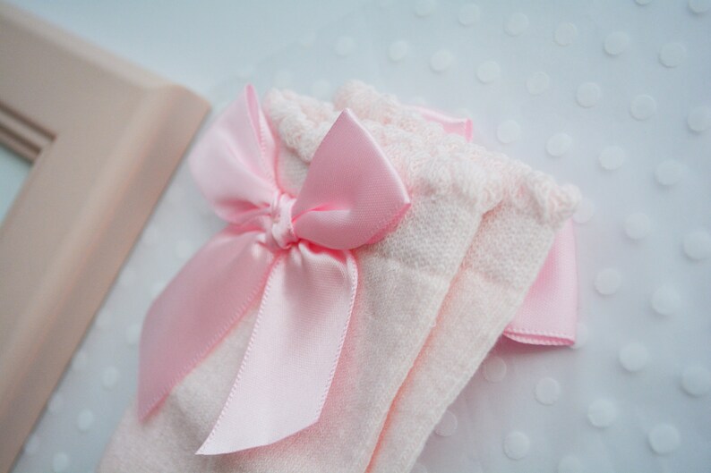Peut inclure: Une paire de chaussettes pour b&eacute;b&eacute; roses avec un n&oelig;ud en satin rose. Les chaussettes sont en tissu doux et tricot&eacute;.