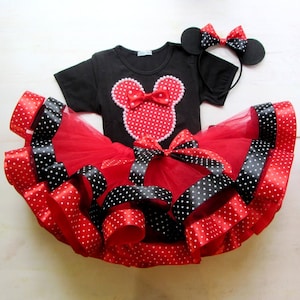 May include: A red and black polka dot tutu skirt with a black t-shirt featuring a red polka dot Minnie Mouse head. The set also includes a black headband with Minnie Mouse ears.