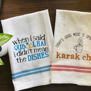 May include: Two white tea towels with printed text. One reads "When I said QUBOOL HAI I didn't mean the DISHES" with blue and gold details. The other says "TODAY'S GOOD MOOD IS SPONSORED BY karak chai" with a coffee cup illustration.
