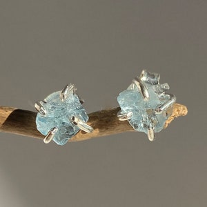 Puede incluir: Un par de pendientes de plata con piedras de aguamarina azul claro. Las piedras son en bruto y tienen una forma natural irregular.