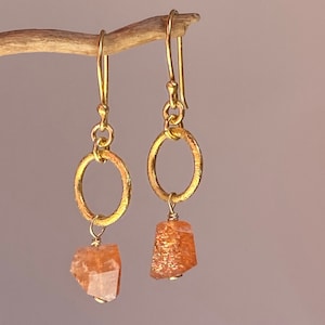 Puede incluir: Pendientes de aro de oro con una piedra de sol naranja colgante.