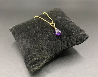May include: A gold chain necklace with a purple gemstone pendant. The pendant is a teardrop shape and is attached to the chain with a small gold ring.