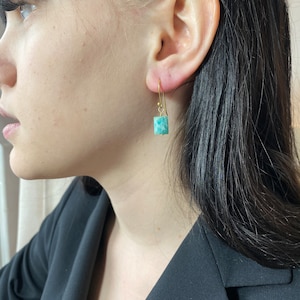 May include: A pair of gold earrings with a square-shaped green gemstone. The earrings are dangling and have a simple design.
