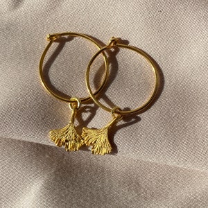 Puede incluir: Pendientes de aro dorados con un dije de hoja de ginkgo colgando de cada aro.