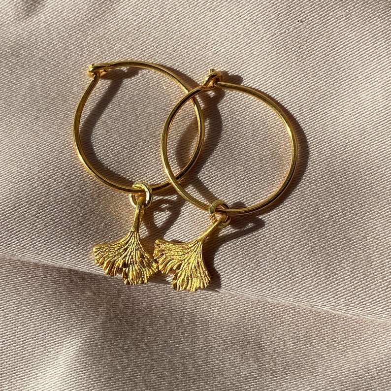 Puede incluir: Pendientes de aro dorados con un dije de hoja de ginkgo colgando de cada aro.