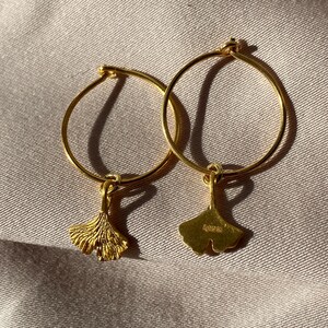 Puede incluir: Un par de pendientes de aro de oro con un dije de hoja de ginkgo colgando de cada aro.