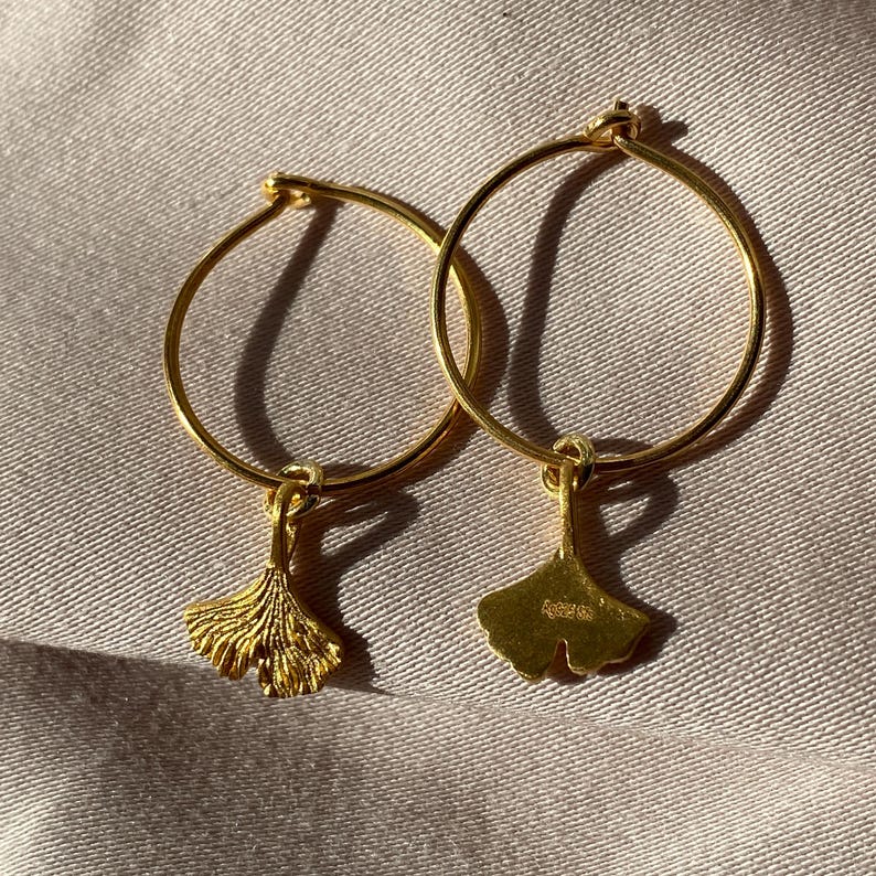 Puede incluir: Un par de pendientes de aro de oro con un dije de hoja de ginkgo colgando de cada aro.