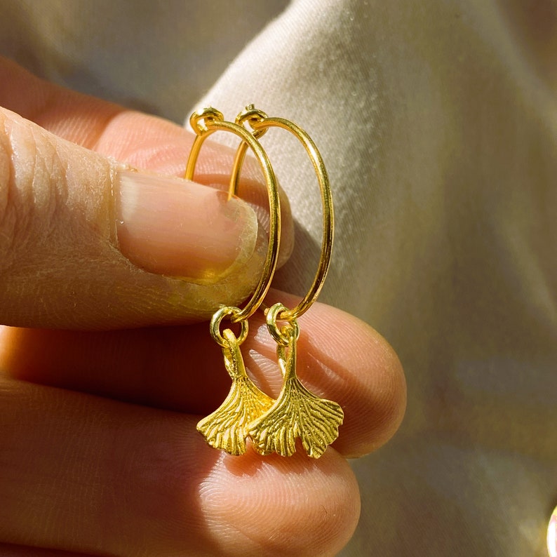 Puede incluir: Un par de pendientes de aro de oro con un dije de hoja de ginkgo colgando de cada aro.