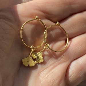 Puede incluir: Un par de pendientes de aro dorados con un dije de hoja de ginkgo. Los pendientes son simples y elegantes, y el dije agrega un toque de naturaleza.