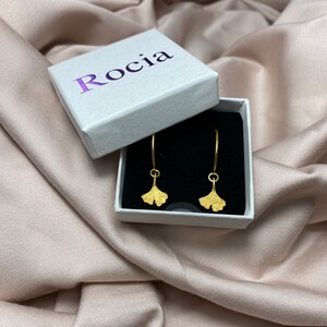 Puede incluir: Pendientes de hoja de ginkgo de oro en una caja blanca con la palabra "Roc&iacute;a" impresa en la tapa.