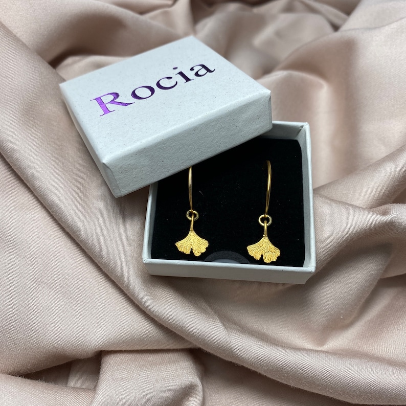 Puede incluir: Pendientes de hoja de ginkgo de oro en una caja blanca con la palabra "Roc&iacute;a" impresa en la tapa.