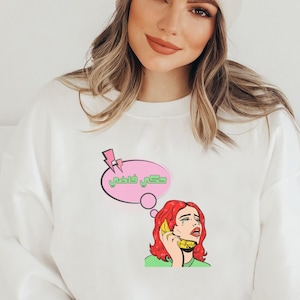 May include: White sweatshirt with a pink speech bubble that says "Just vibing" in green text. The speech bubble is above a cartoon image of a woman with red hair talking on a yellow phone.