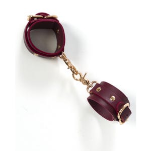 Puede incluir: Un par de esposas de cuero burdeos con herrajes dorados. Las esposas están conectadas por una cadena.