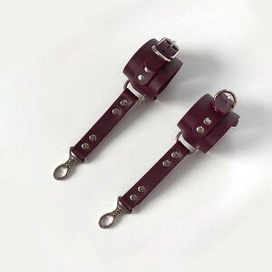 Puede incluir: Par de esposas de cuero burdeos con herrajes plateados. Cada esposa tiene un brazalete con hebilla, una correa con remaches y un mosquetón. Se muestran sobre un fondo blanco.
