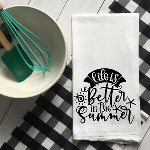 Peut inclure: Torchon de cuisine blanc avec l'inscription "Life is Better in the Summer" en noir. Le torchon est à côté d'un bol blanc, d'une spatule verte et d'un fouet. Un tissu à carreaux noir et blanc est en arrière-plan.