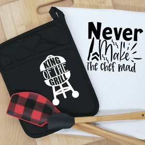 Puede incluir: Un guante de cocina negro con un gráfico blanco de una parrilla y el texto "KING OF THE GRILL". Una espátula a cuadros rojos y negros, un paño de cocina blanco con el texto "Never Make the chef mad" y dos utensilios de cocina de madera.