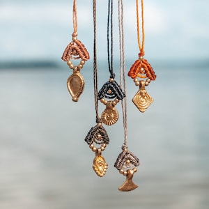 Könnte beinhalten: Eine Sammlung handgefertigter Halsketten mit aufwendigen Makramee-Details und goldfarbenen Anhängern. Die Anhänger weisen verschiedene Formen auf, darunter Tropfen, geometrische Designs und abstrakte Formen. Die Halsketten hängen vor einem verschwommenen Hintergrund aus Wasser und Himmel.