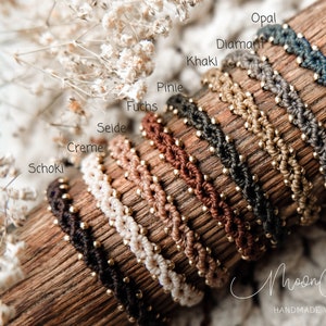 May include: A close-up of ten colorful braided bracelets with gold beads. The bracelets are arranged in a row on a wooden surface. The colors of the bracelets are:  Schoki, Creme, Seidel, Fuchs, Pinie, Khaki, Diamant, Opal.