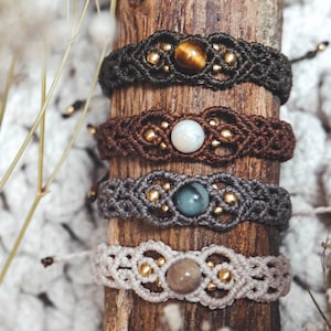 May include: Four macrame bracelets featuring central round gemstones in tiger eye, white, blue-gray, and beige. Each bracelet is crafted with knotted cord and small metallic gold beads arranged in a symmetrical pattern around the center stone.