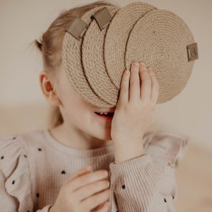以下が含まれることがあります： 茶色の髪の若い人が、顔の前に4枚の織り込まれた茶色のコースターをセットで持っています。コースターは自然素材で作られており、素朴な外観をしています。