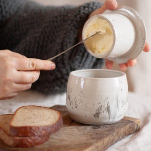 Puede incluir: Una mantequera de cerámica blanca con tapa, que se utiliza para sacar mantequilla. El plato está junto a una tabla de cortar de madera con rebanadas de pan y pequeños tomates rojos. La mantequera tiene un diseño rústico y moteado.