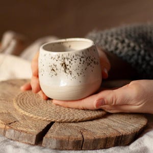 Puede incluir: Una taza de cerámica hecha a mano con un diseño moteado. La taza es blanca con motas marrones y una base beige claro. Se sostiene sobre un posavasos de madera y una esterilla tejida.