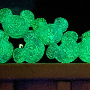 May include: A group of six green glow-in-the-dark Mickey Mouse head shaped pumpkins. Each pumpkin has a carved jack-o'-lantern face with a toothy grin.