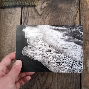 Könnte beinhalten: Schwarz-Weiß-Fotografie von Meereswellen, die an einer dunklen Küste brechen. Das Bild wird vor einem Holzgrund gehalten, mit Stoffmustern in der oberen linken Ecke.