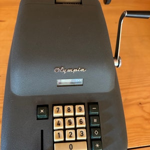 Puede incluir: Una máquina de sumar Olympia vintage gris con una manija negra y un teclado numérico blanco. La máquina tiene una ventana de visualización negra y una palanca negra en el lado izquierdo.
