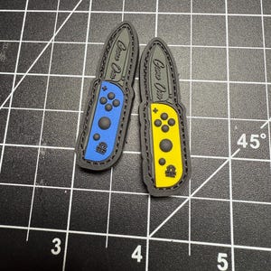 May include: Two patches shaped like game controller joysticks. One is blue, the other yellow, both with black trim and the words "Game Over" printed on them. Each has a small emblem at the bottom.