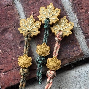 Puede incluir: Tres tiradores de cremallera con dijes de hoja de arce color amarillo mostaza hechos de material moldeado con detalles de venas en relieve. Cada tirador está unido a un cordón paracord estampado en tonos verde, marrón y naranja con un nudo.