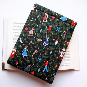 May include: A rectangular fabric book sleeve featuring a black background with a repeating pattern of nutcrackers, ballerinas, candy canes, Christmas ornaments, and green pine branches. The sleeve is folded over at the top edge.