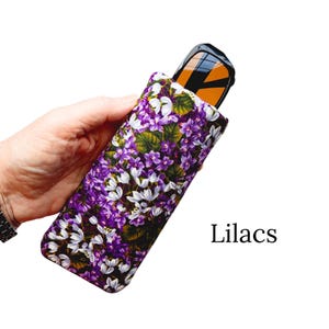 Pu&ograve; includere: Un astuccio floreale per occhiali con un motivo di lilla viola e bianco. L'astuccio contiene un paio di occhiali da sole con lenti arancioni. La parola "Lilacs" &egrave; stampata in nero sul lato destro dell'immagine.