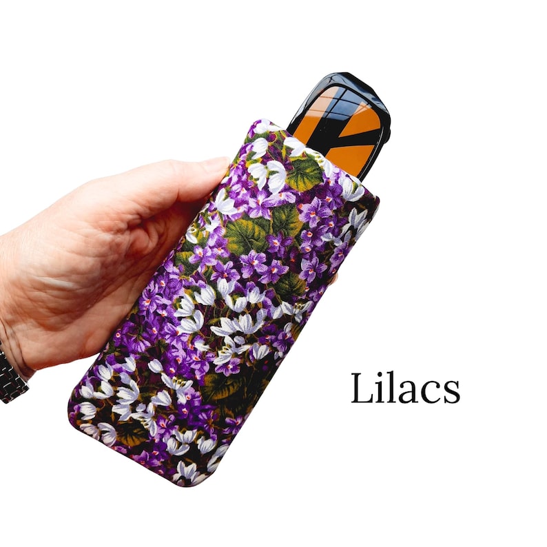 Pu&ograve; includere: Un astuccio floreale per occhiali con un motivo di lilla viola e bianco. L'astuccio contiene un paio di occhiali da sole con lenti arancioni. La parola "Lilacs" &egrave; stampata in nero sul lato destro dell'immagine.