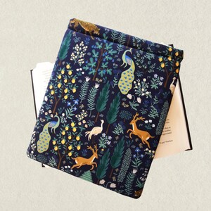 May include: A navy blue book sleeve with a colorful forest pattern. The design includes peacocks, deer, trees, and other foliage. The book sleeve is open, revealing the pages of a book.