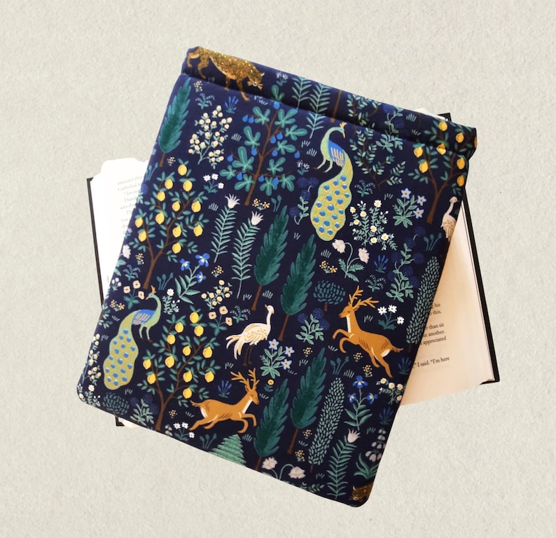 May include: A navy blue book sleeve with a colorful forest pattern. The design includes peacocks, deer, trees, and other foliage. The book sleeve is open, revealing the pages of a book.