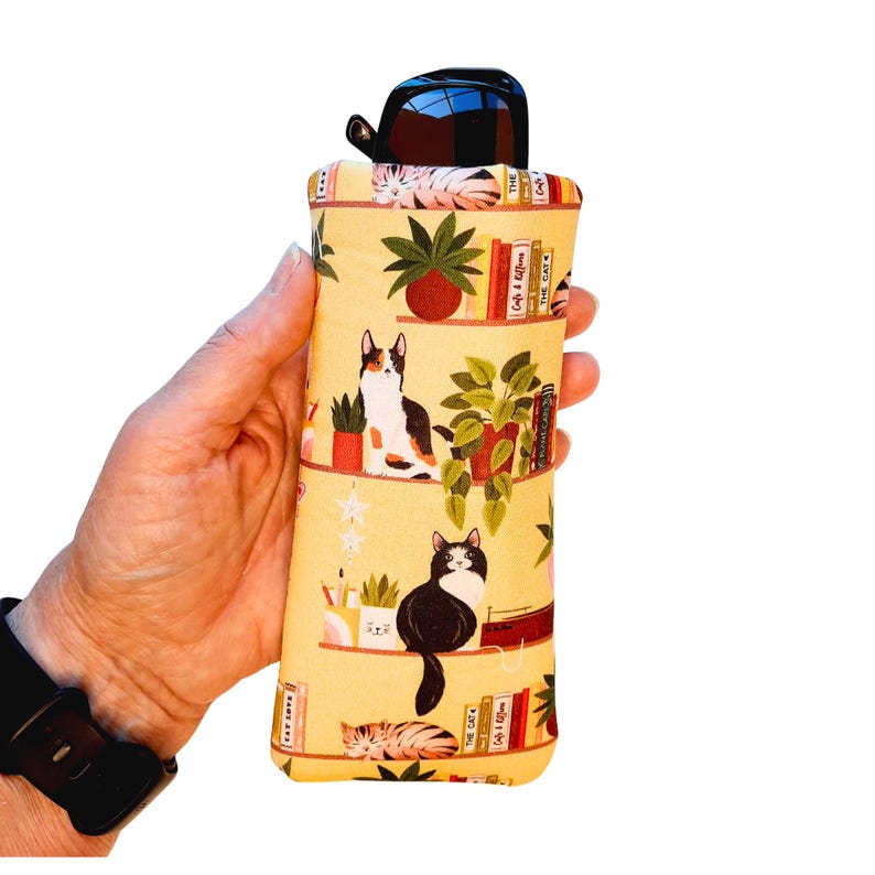 Pu&ograve; includere: Un astuccio giallo per occhiali con un motivo a gatti e librerie. L'astuccio contiene un paio di occhiali da sole neri. Il design presenta gatti, piante in vaso e libri sugli scaffali. L'astuccio &egrave; tenuto in mano.