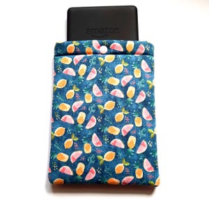 May include: A blue fabric sleeve with a fruit pattern, holding a black electronic device with the Amazon logo. The sleeve features a button closure and is decorated with orange and pink fruit slices and green leaves.