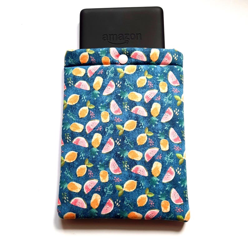 May include: A blue fabric sleeve with a fruit pattern, holding a black electronic device with the Amazon logo. The sleeve features a button closure and is decorated with orange and pink fruit slices and green leaves.
