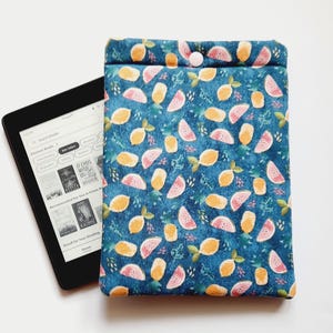 May include: A blue fabric e-reader case with a pattern of lemons and watermelon slices. The case has a button closure and is next to a black e-reader displaying a book selection screen.