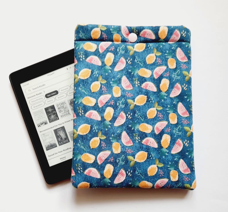 May include: A blue fabric e-reader case with a pattern of lemons and watermelon slices. The case has a button closure and is next to a black e-reader displaying a book selection screen.