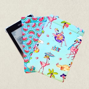 May include: Kindle e-reader with two fabric cases. One case has a watermelon pattern, the other has a flamingo design with palm trees, beach scenes, and a flamingo in a float. The cases are blue, pink, and green.