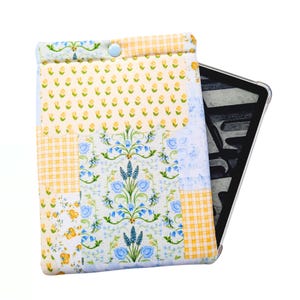 May include: A quilted e-reader case with a patchwork design. The case features a variety of floral and gingham patterns in yellow, blue, and white. A light blue button secures the top flap. The case is designed to hold an e-reader.